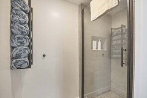 Ein modernes Badezimmer mit einer Duschkabine aus Glas, einem Handtuchhalter mit gefalteten HandtĂŒchern und einer kleinen Ablage mit Pflegeprodukten.