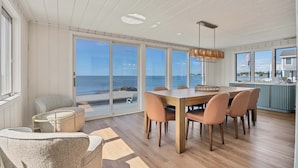 A dining area with a wooden table and chairs, large windows with a view of the beach, and a hanging light fixture.
