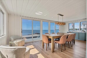 Un coin repas avec une table et des chaises en bois, de grandes fenĂȘtres donnant sur la plage et un luminaire suspendu.