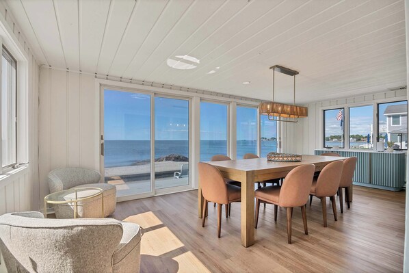 A dining area with a wooden table and chairs, large windows with a view of the beach, and a hanging light fixture.
