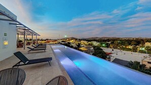 Une piscine à débordement avec des chaises longues, un espace salon abrité et une vue sur le paysage urbain au crépuscule.