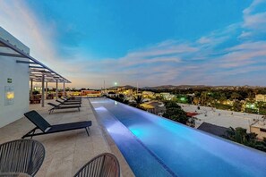 An infinity pool with lounge chairs, a covered seating area, and a view of a cityscape at dusk.