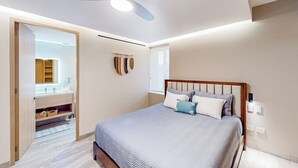 Une chambre à coucher avec un lit, un ventilateur de plafond, une table de chevet et une salle de bain visible à travers une porte ouverte.
