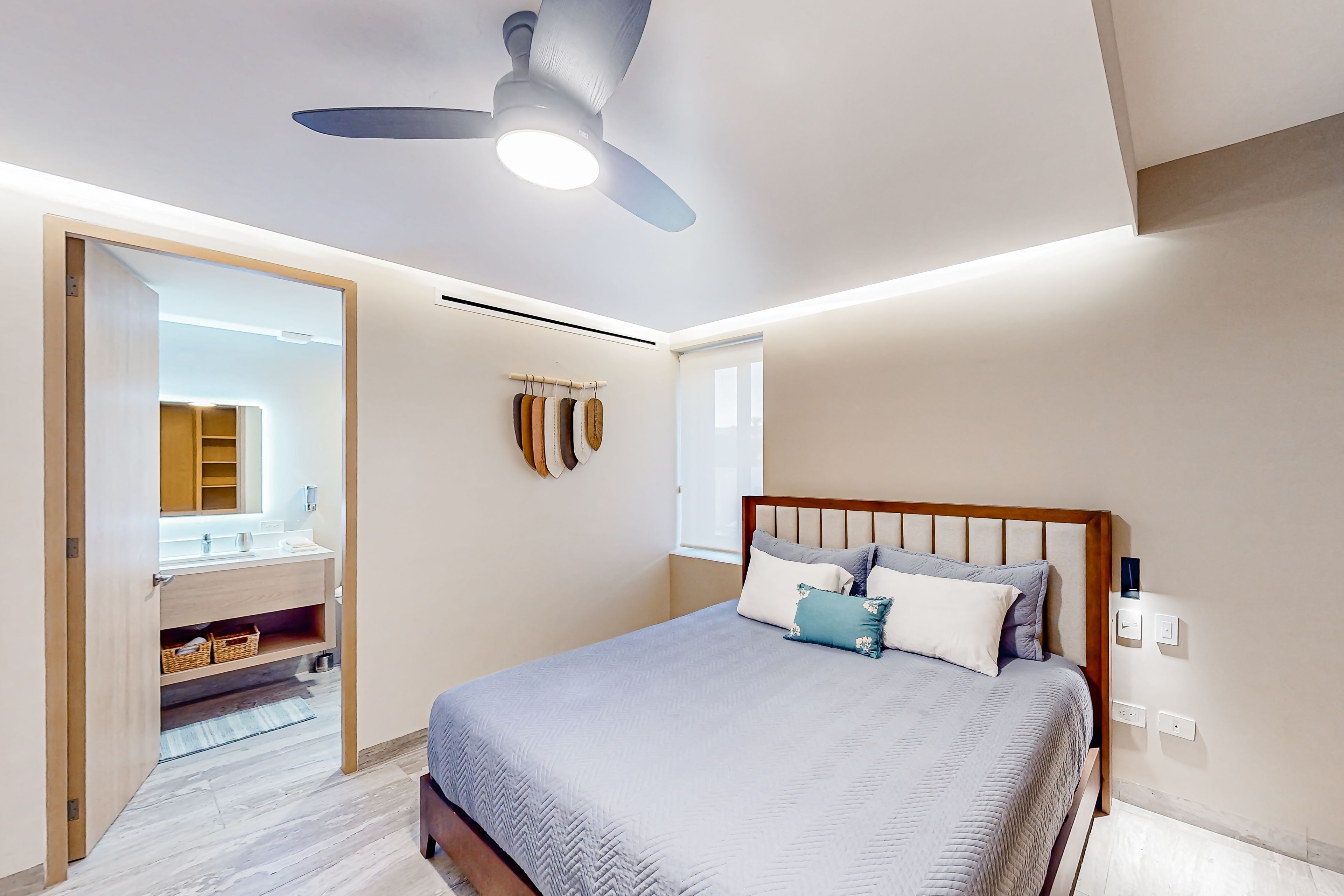 Une chambre à coucher avec un lit, un ventilateur de plafond, une fenêtre et une salle de bain visible à travers une porte ouverte.