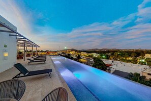 An infinity pool with lounge chairs and a covered seating area, overlooking a cityscape at dusk. - Downtown Condo With Great Views, Shared Pool, Hot Tub, & Grill (San José del Cabo)