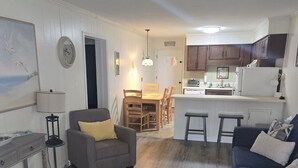 Uma sala de estar moderna com um sofá cinza, um sofá azul, uma área de refeições com mesa de madeira e uma cozinha com armários de madeira e uma geladeira branca.