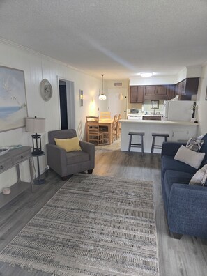 Ein modernes Wohnzimmer mit einem grauen Sofa, einem blauen Sofa, einem Essbereich mit einem Holztisch und einer Küche mit Holzschränken und einem weißen Kühlschrank.