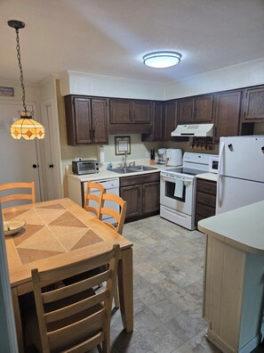 Una cocina con armarios de madera, una mesa de comedor con sillas, un refrigerador, una estufa y una cafetera.