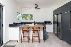 Eine moderne Küche mit einer zentralen Kochinsel, zwei hölzernen Barhockern, einem Edelstahlkühlschrank und einem Deckenventilator.
