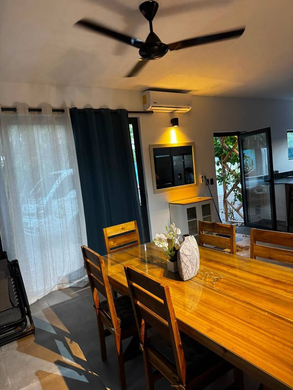 A dining area with a wooden table, chairs, a ceiling fan, an air conditioning unit, and a view of the outdoors.
