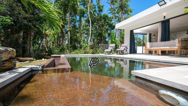 Une piscine moderne à l’eau cristalline, entourée d’une végétation luxuriante et d’un bâtiment contemporain doté d’un espace de détente extérieur.