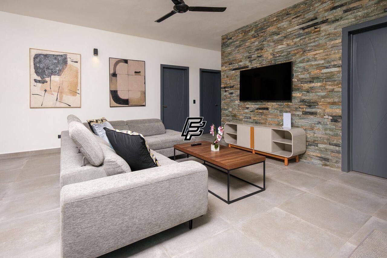 A modern living room with a grey sofa, a coffee table, a flat-screen TV mounted on a stone wall, and a ceiling fan.