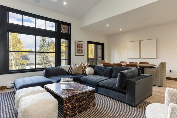 A modern living room with a sectional sofa, a marble coffee table, and large windows with a view of trees. - Modern Mountain Home with Lux Kitchen & Cozy Living Room Heart of Beaver Creek (Beaver Creek)