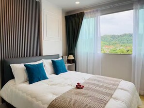 Habitación de hotel con cama, mesita de noche, lámpara y ventana con vistas a zonas verdes.