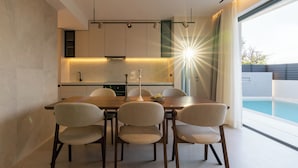 A modern dining area with a wooden table and chairs, a kitchen in the background, and a view of a swimming pool. - Green villas, perfect for relaxing family or friends’ vacations (Rethymno)