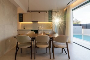 A modern dining area with a wooden table and chairs, a kitchen in the background, and a view of a swimming pool. - Green villas, perfect for relaxing family or friends’ vacations (Rethymno)