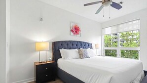 Ein Schlafzimmer mit Bett, Nachttischen, Deckenventilator, Fenster mit Blick und einem an der Wand hängenden Kunstwerk.