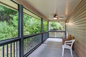 A covered porch with a ceiling fan, wooden railing, and a view of dense greenery.