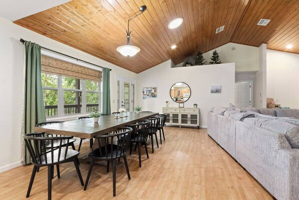 A modern kitchen with a wooden ceiling, a dining area with a long table and chairs, and a grey sofa.