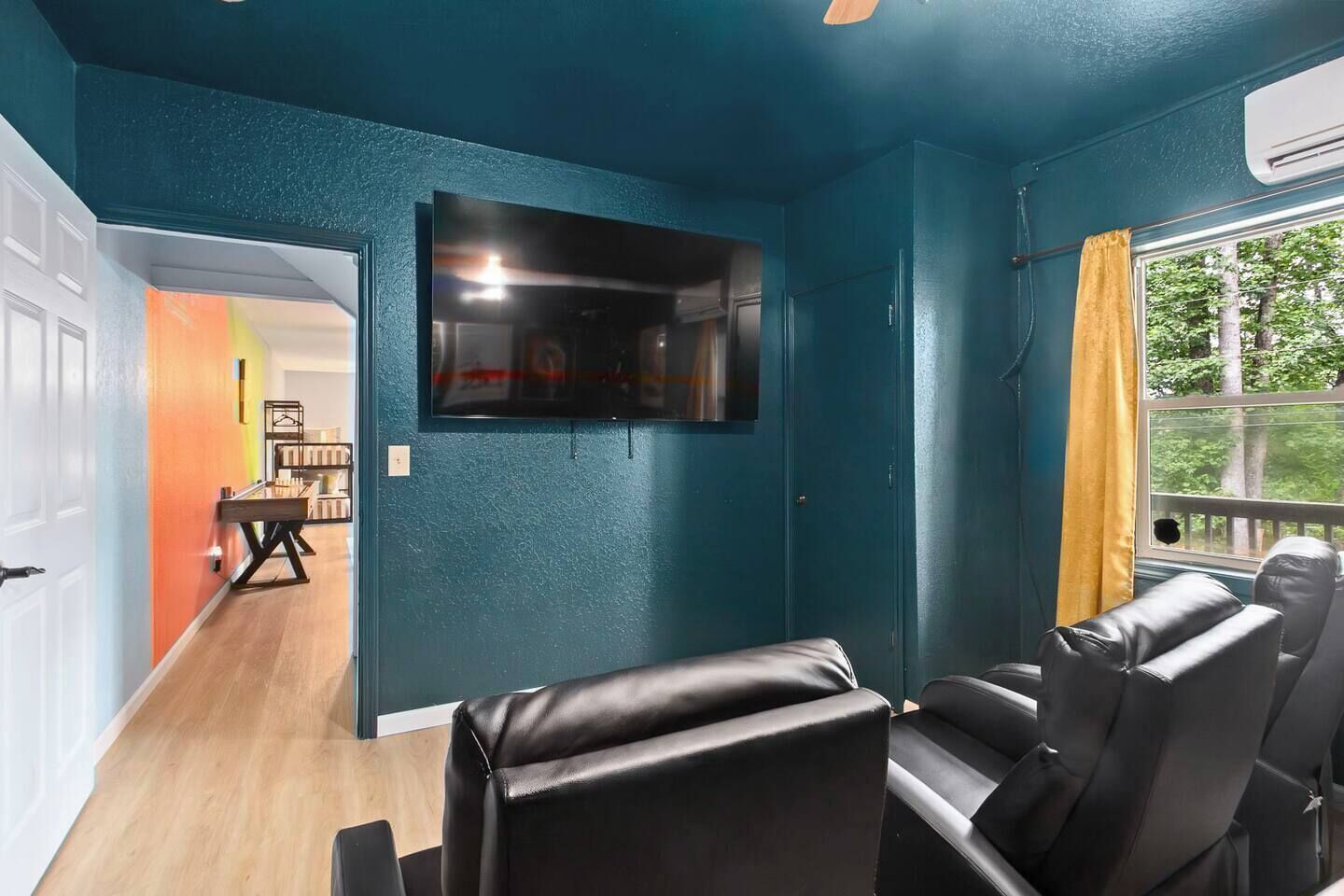 Ein Zimmer mit türkisfarbenen Wänden, einem an der Wand befestigten Fernseher, Ledersitzmöbeln und einem Fenster mit Blick auf Bäume.