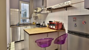 Een compacte keuken met een fornuis, broodrooster en koelkast, een houten aanrecht en twee paarse barkrukken.