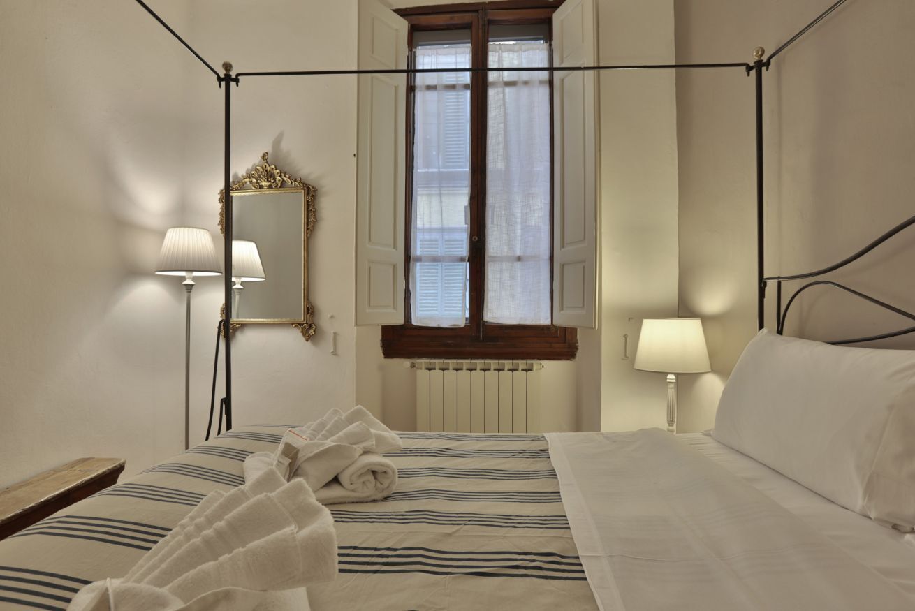 A hotel room with a four-poster bed, two bedside lamps, a mirror, a window with shutters, and a radiator.