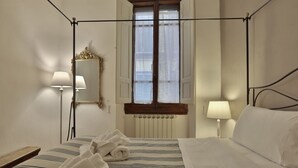 Een hotelkamer met een hemelbed, twee bedlampen, een spiegel, een raam met shutters en een radiator.