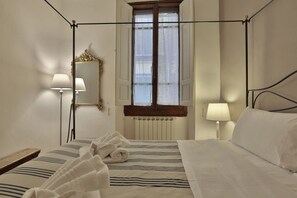 Ein Hotelzimmer mit Himmelbett, zwei Nachttischlampen, einem Spiegel, einem Fenster mit Rollläden und einer Heizung.