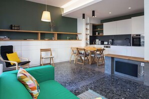 Eine moderne Küche mit weißen Schränken, einem Essbereich mit Holzsesseln, einem grünen Sofa und einem an der Wand montierten Fernseher.
