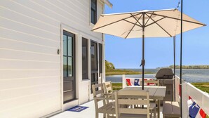 A deck with a table and chairs, a grill, and a view of the water. - Edgewater Sanctuary | Fully Off Grid Ocean Cottage | Directly on Buzzard's Bay (Wareham)