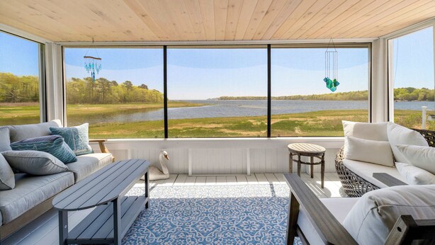 A spacious room with a large window overlooking a lake, a wooden ceiling, and a seating area with a bench and cushions. - Edgewater Sanctuary | Fully Off Grid Ocean Cottage | Directly on Buzzard's Bay (Wareham)