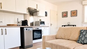 A modern kitchen with white cabinets, a microwave, an oven, and a coffee maker. There's a beige sofa with a patterned pillow and a small table with two blue mugs. - Cocoon apartment in Sanguinet - lake, forest, Pilat dune and ocean (Sanguinet)