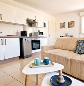 Eine moderne Küche mit weißen Schränken, einer Mikrowelle, einem Backofen und einer Kaffeemaschine. Ein beiges Sofa mit einem gemusterten Kissen und ein kleiner Tisch mit zwei blauen Tassen.