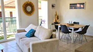 A living room with a sofa, a dining table with chairs, a window with a view, and a wall-mounted decorative item. - Cocoon apartment in Sanguinet - lake, forest, Pilat dune and ocean (Sanguinet)