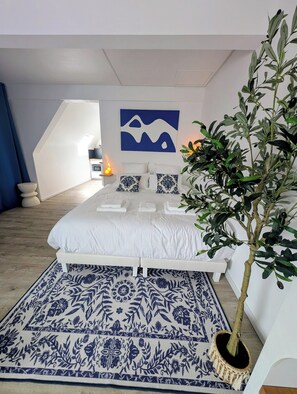 Une chambre à coucher avec un lit, des oreillers, un tapis à motifs bleus et blancs, un tableau abstrait accroché au mur, une plante en pot et une table de chevet avec une lampe.