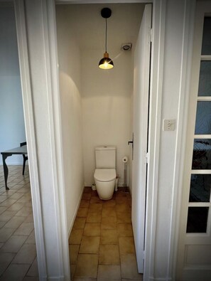 A narrow hallway leading to a bathroom with a toilet, a hanging light fixture, and a tiled floor. - Vrbo Property (Arles)