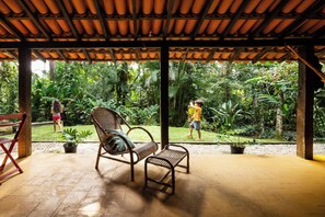 Un espacio al aire libre techado con piso de madera, dos sillas de mimbre y vista a vegetación exuberante.