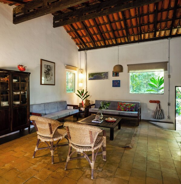 Sala de estar con techo alto de madera, un sofá, dos sillones de mimbre, una mesa de centro, un aparador y una ventana con vistas a zonas verdes.