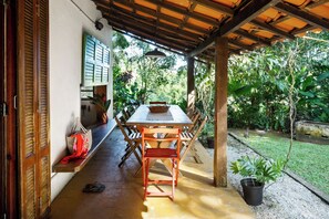 En udendørs overdækket terrasse med et træbord og stole, omgivet af grøn vegetation.