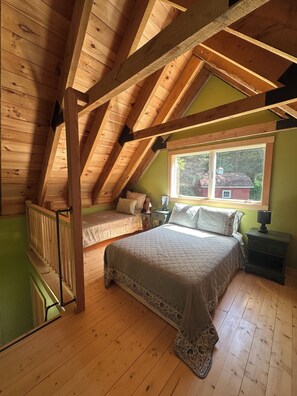 A loft bedroom with a sloped ceiling, a large bed, a window with a view of a red house, and a wooden floor.