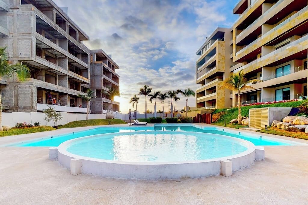 Une piscine entourée d’immeubles à plusieurs étages dotés de balcons, de palmiers et surplombant un ciel dégagé.