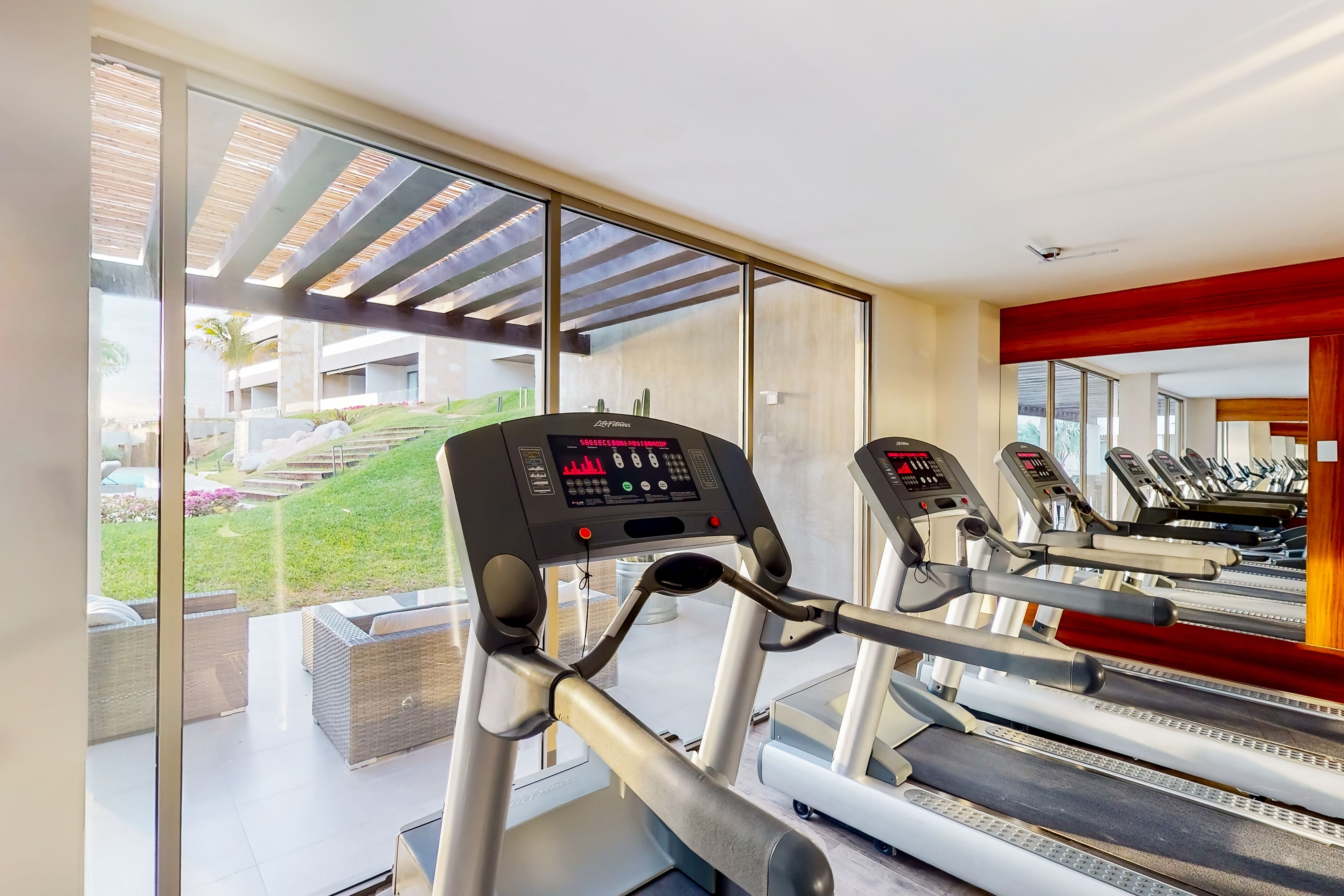 Une salle de sport équipée de tapis de course et d’autres appareils d’exercice, dotée de grandes fenêtres et offrant une vue sur un espace paysager extérieur.