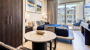 A modern hotel room with a bed, a round table, a sofa, and a view of the city through large windows. - Exquisite Apartment With Balcony, TV, Wifi, Heated Pool & Gym/fitness Room (Cabo San Lucas)