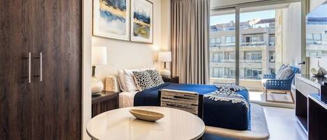 Ein modernes Hotelzimmer mit Bett, rundem Tisch, Sofa und Blick auf die Stadt durch große Fenster.