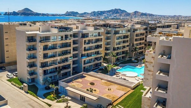 A high-rise hotel with multiple balconies, a swimming pool, and a view of the sea and mountains. - Exquisite Apartment With Balcony, TV, Wifi, Heated Pool & Gym/fitness Room (Cabo San Lucas)