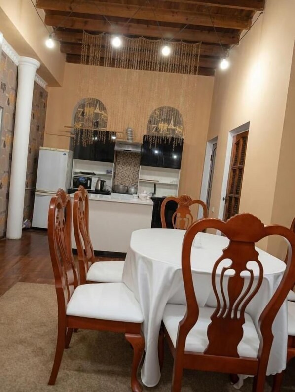 A dining area with wooden chairs and a table, a kitchen in the background, and a hanging light fixture. - Ayvan Eco Hostel (Tashkent)