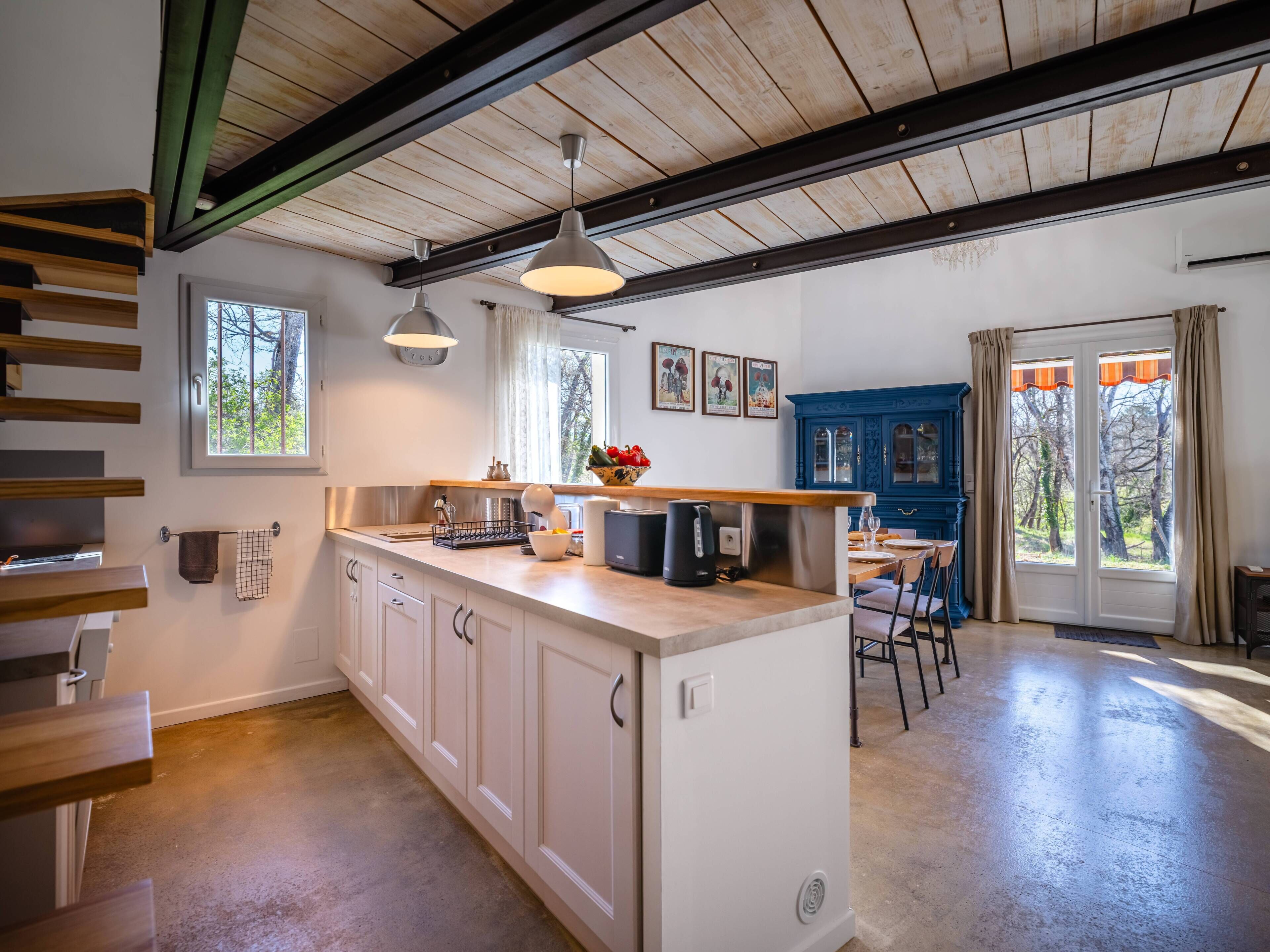 A kitchen with wooden beams, a central island, pendant lights, and a dining area with a blue cabinet and a view through the glass door.
