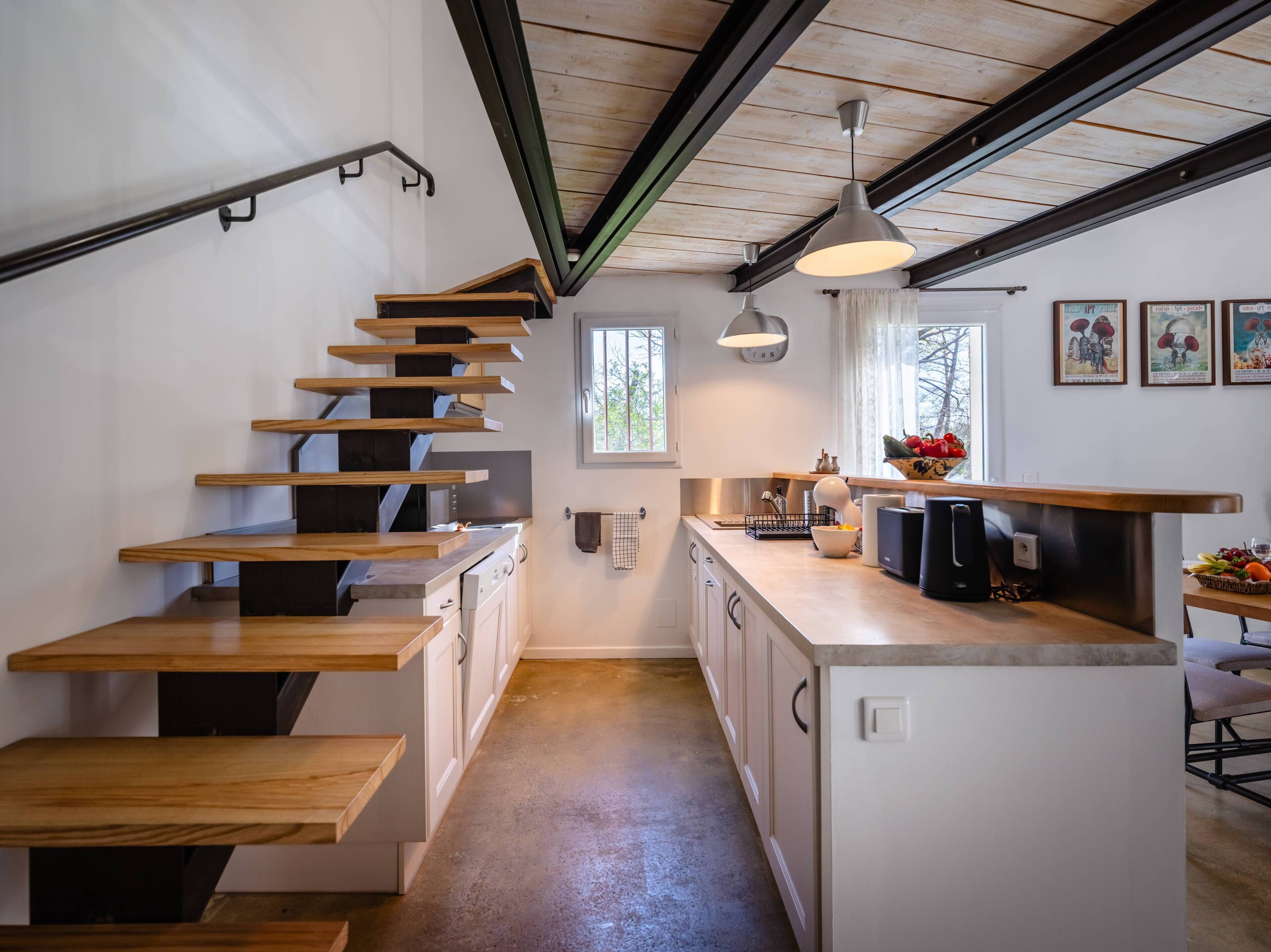 A modern kitchen with a wooden staircase, a central island, and a window providing natural light.