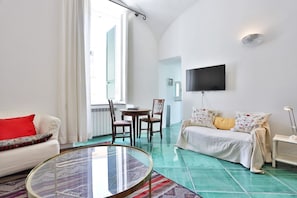 A modern living room with a round glass coffee table, a white sofa with a red pillow, a beige sofa with patterned cushions, a flat-screen TV mounted on the wall, and a large window with white curtains. - Spacious apartment in the heart of Petraio, one of Naples' most authentic neighborhoods, just a 10-minute walk from the Vanvitelli metro station and the Petraio funicular. Located in a 19th-century building, it retains vaulted ceilings, fine details and  (Napoli)