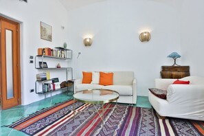 Ein Wohnzimmer mit einem weißen Sofa, einem runden Glastisch, einem gemusterten Teppich, einem Bücherregal und zwei Wandleuchten.